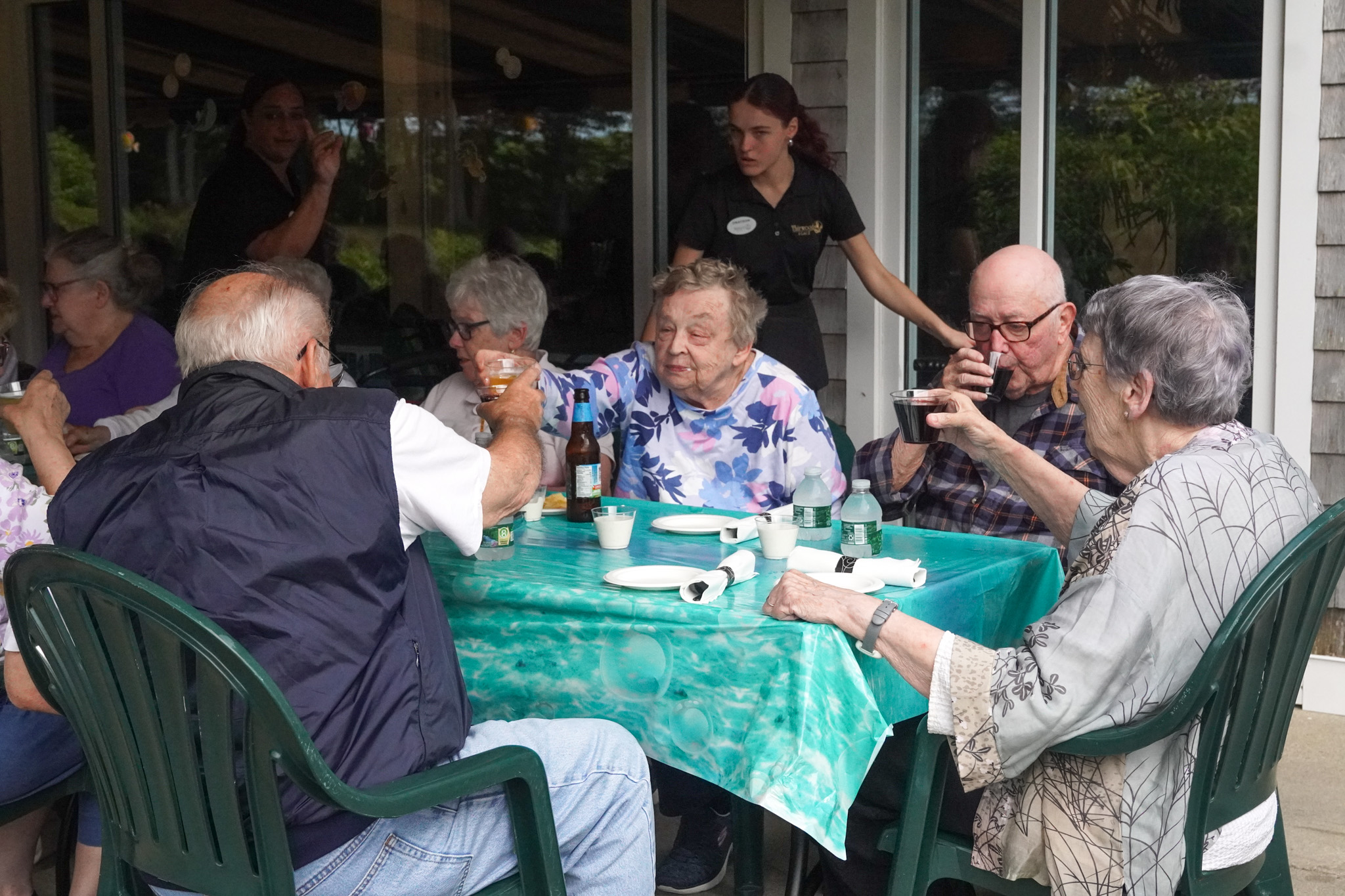 Elderly people sit around a table outdoors, enjoying a summer gathering as they eat, drink, and socialize.