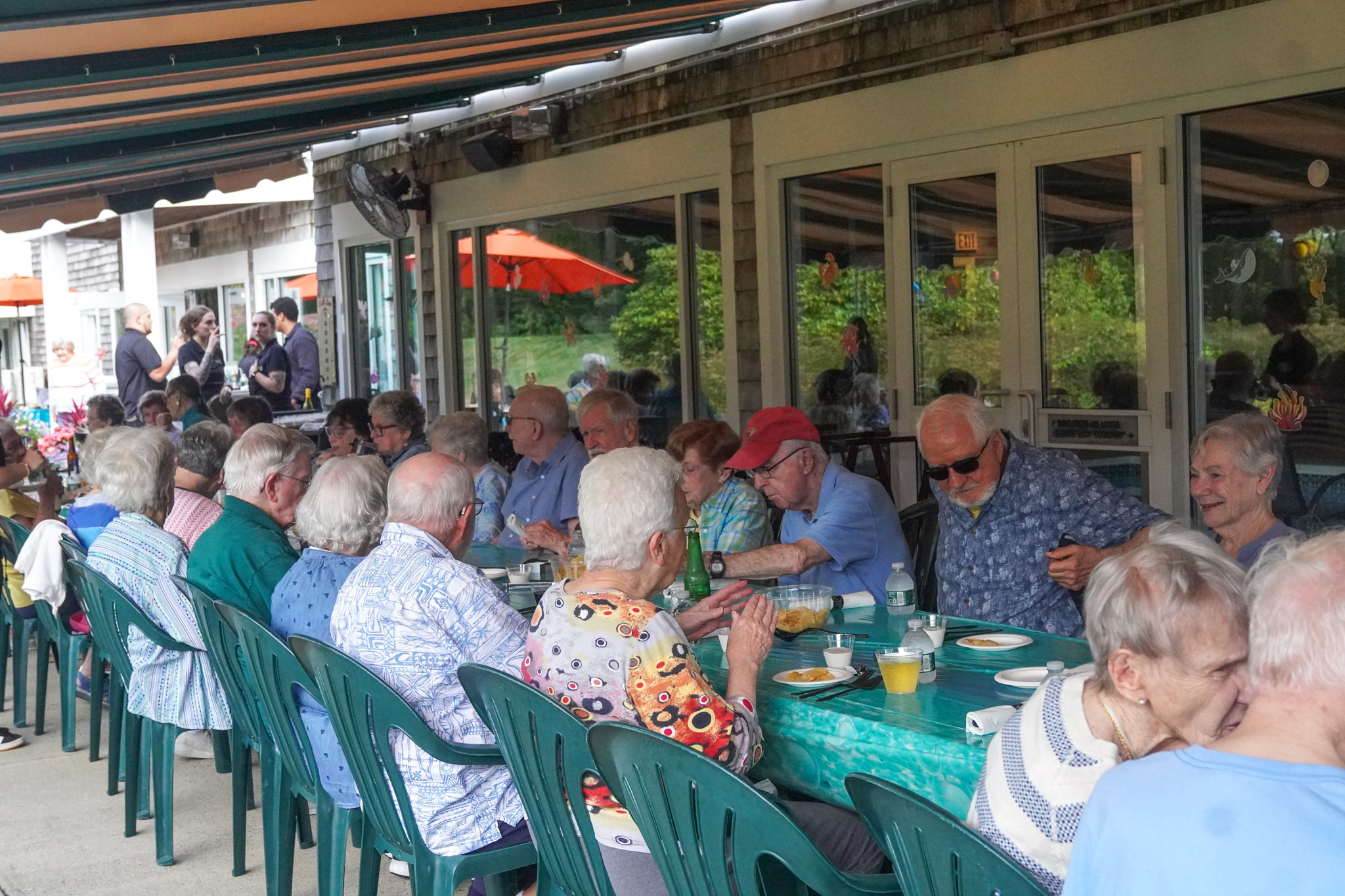 A group of seniors sit at long tables outdoors, enjoying a summer gathering as they eat and socialize in the warm weather.