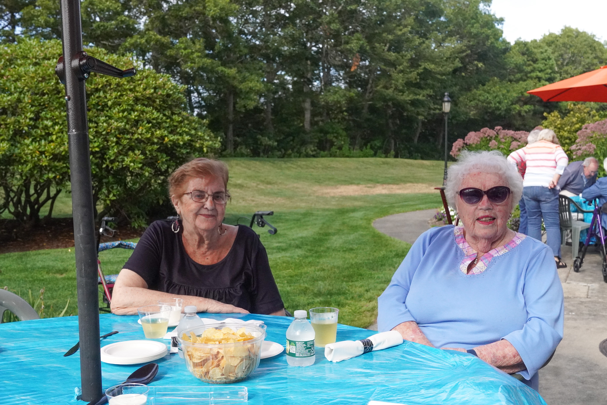 Two older women sit at a table outdoors with snacks and drinks, smiling and enjoying a sunny summer day.