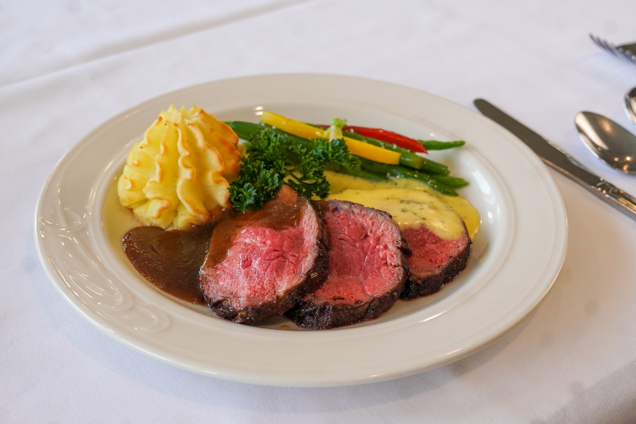 Sliced roast beef with gravy, mashed potatoes, vegetables, and sauce on a white plate.