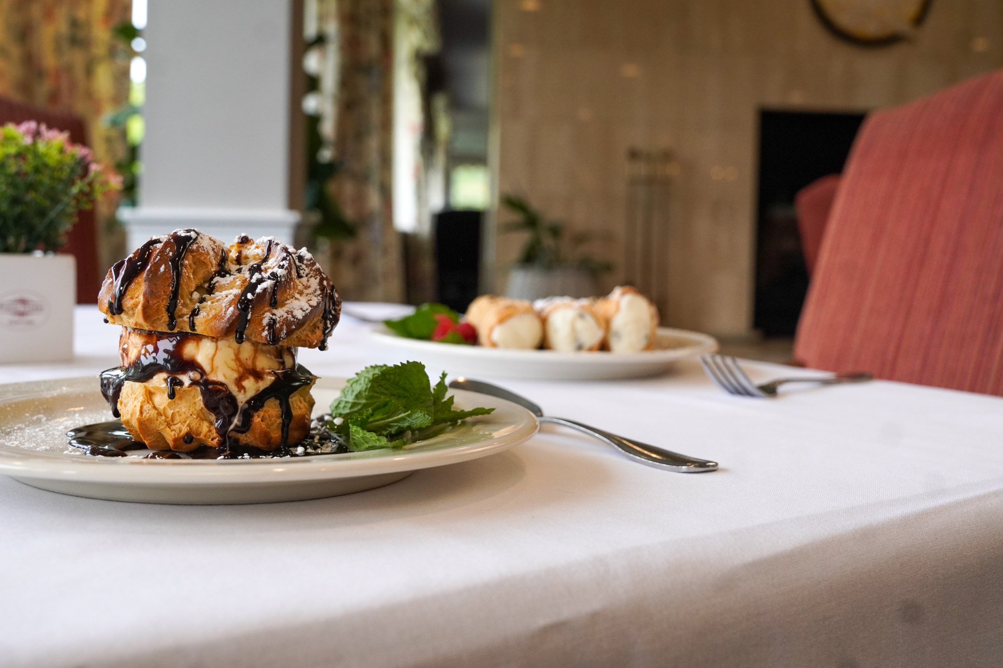 A cream puff dessert drizzled with chocolate sits on a plate, perfect for a birthday treat, with another dessert blurred in the background.