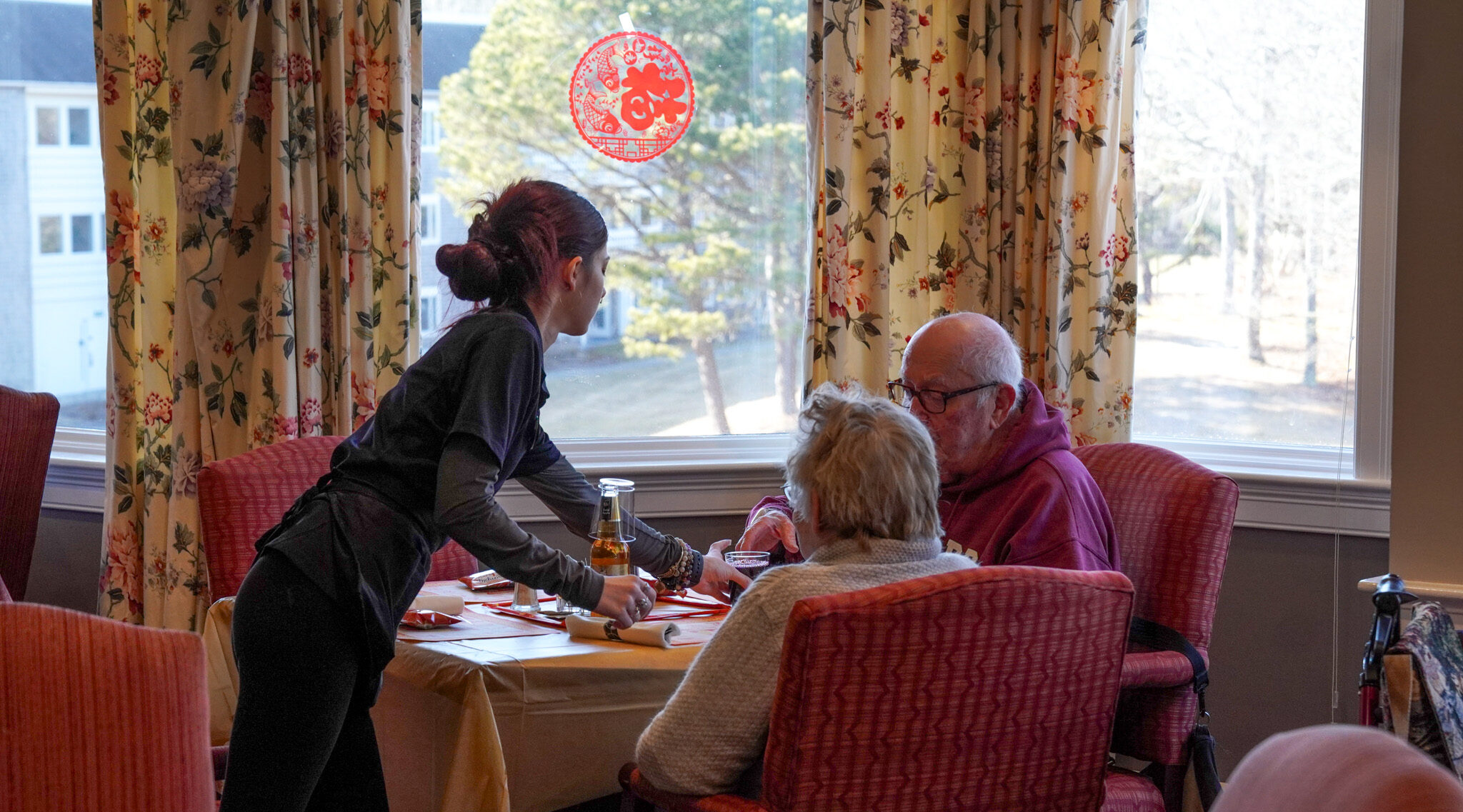 The server assists the elderly couple, who just started downsizing their lives, at a restaurant table by a window adorned with floral curtains.