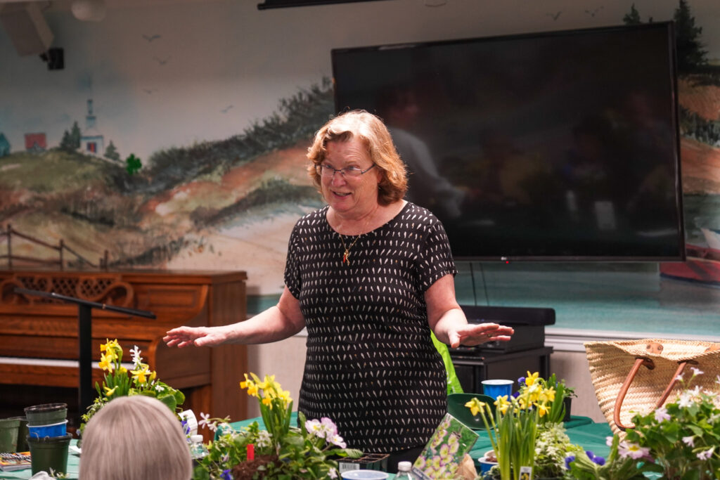 A woman is speaking to a group in a room adorned with a mural, surrounded by potted plants and a large screen behind her. The gathering focuses on senior living, fostering an engaging environment for all attendees.