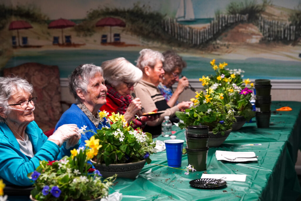 Elderly women sitting at a table in a senior living community, painting and arranging flowers in a room with a beach mural.