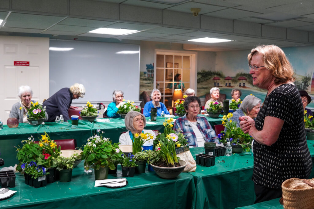 A woman leads a flower arrangement class in a senior living community, guiding seniors seated at tables adorned with green tablecloths and fresh blooms.