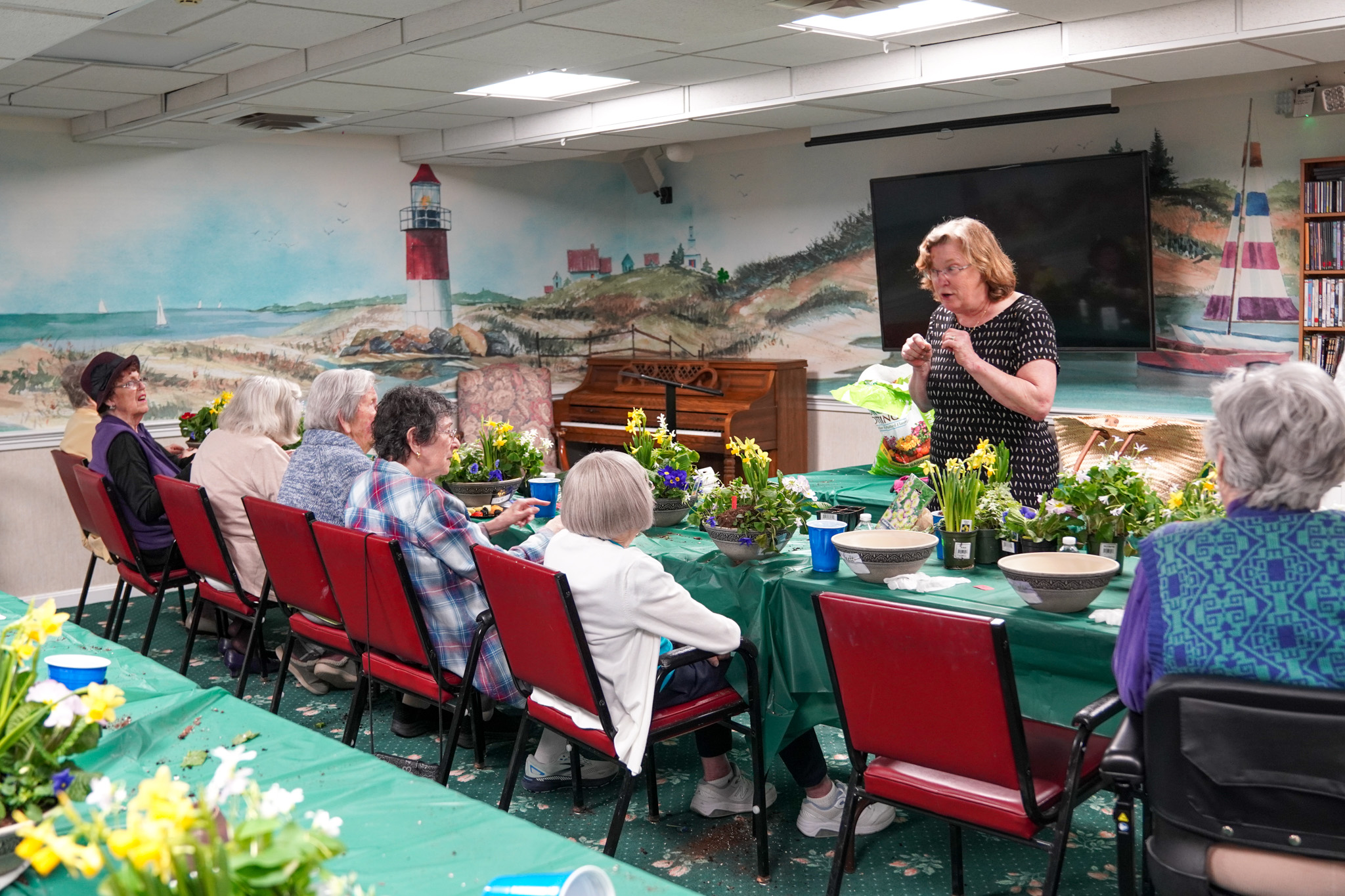 In a cozy room at the senior living center, a group of elderly people gather, attentively listening to a woman share her insights on the art of flower arranging.
