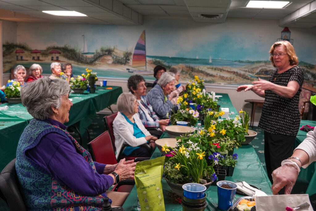 A group of seniors in a senior living community room engage in a delightful flower arranging activity, guided by a cheerful woman standing at the front.