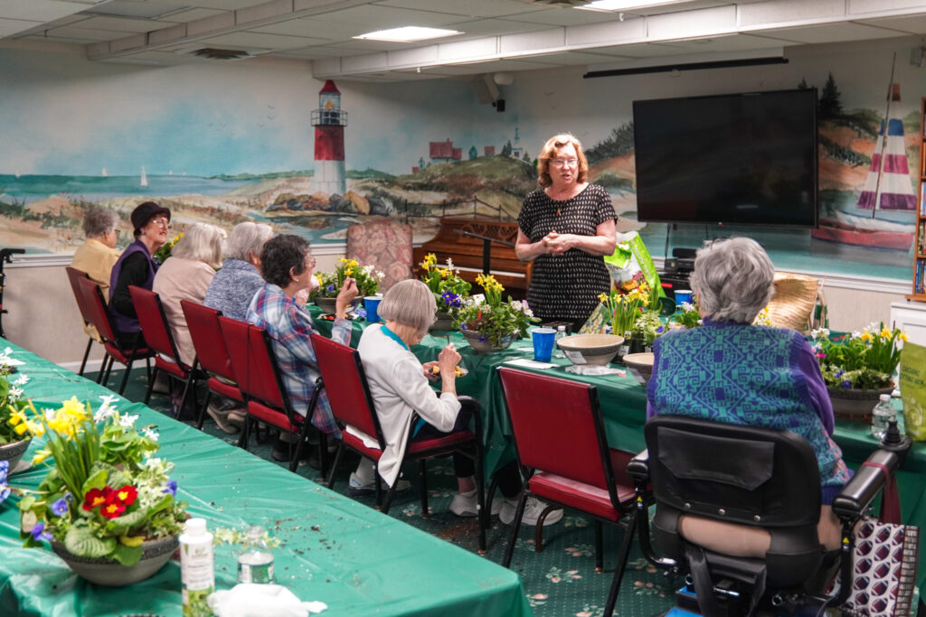 A woman stands speaking in front of a group seated at a table with plants, surrounded by beach-themed murals, creating a serene atmosphere perfect for discussing senior living.