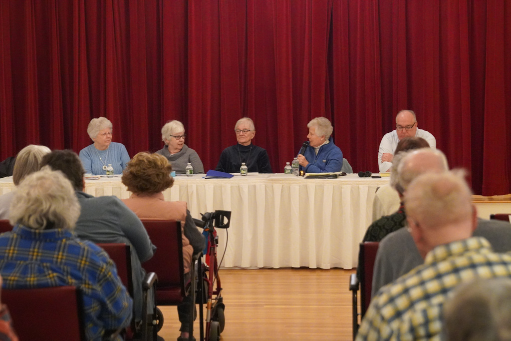 Five people sit at a panel table with microphones, discussing scholarship opportunities, as they face an audience in a room with red curtains.