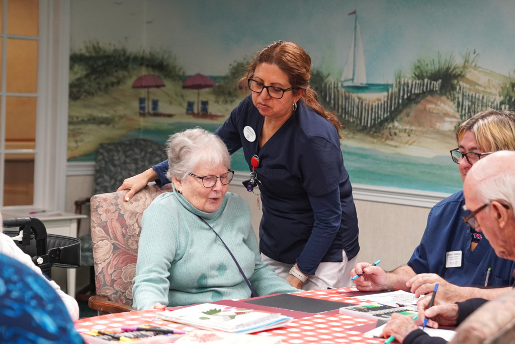 A caregiver assists an elderly woman at a table during an arts and crafts activity, fostering a sense of community in the care facility.