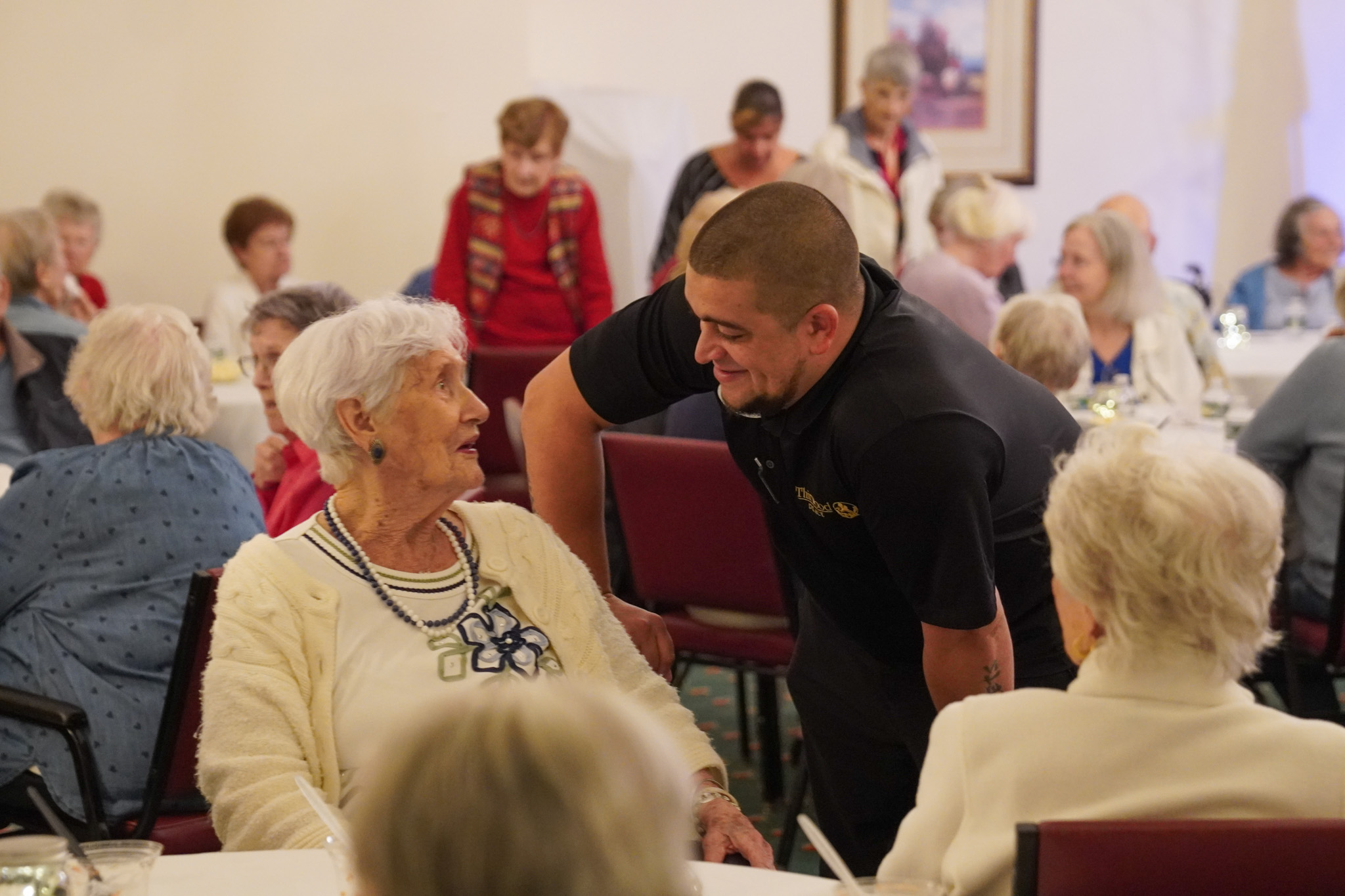A man smiles and talks with an elderly woman at a busy gathering of seniors in a community room, discussing the impact of a local scholarship program.