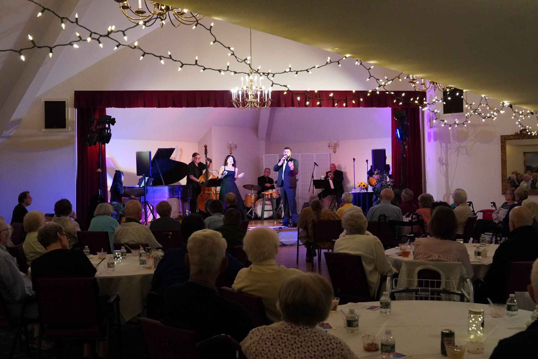 A jazz band performs on stage for an audience in a decorated hall with string lights and chandeliers, celebrating the recipients of a prestigious scholarship.