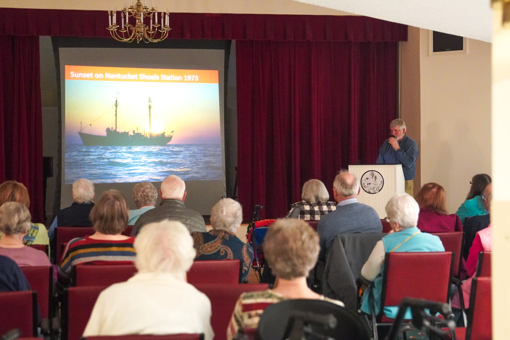 A group, including a nurse, watches a man present a slideshow of a sunset over a ship at a Nantucket Shoals station.