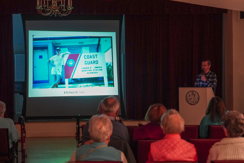 A man gives a presentation to an audience with a Coast Guard image projected on a screen behind him, while describing the vital teamwork between coast guards and nurse professionals during emergencies.