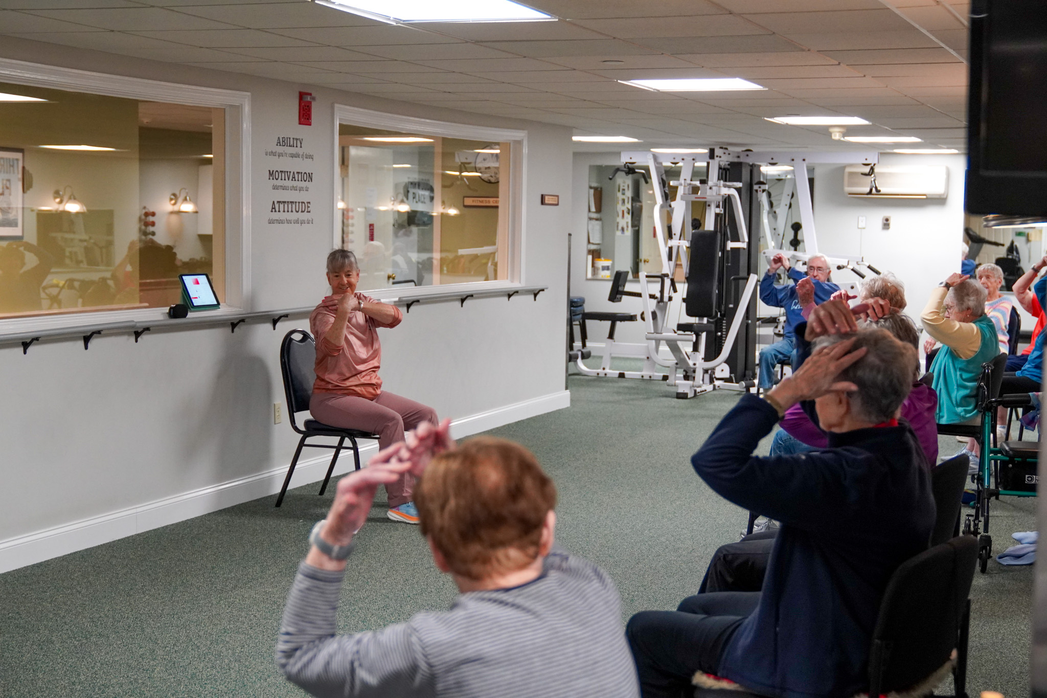 In a downsized gym setting, a group fitness class for seniors features seated participants following an instructors guidance.