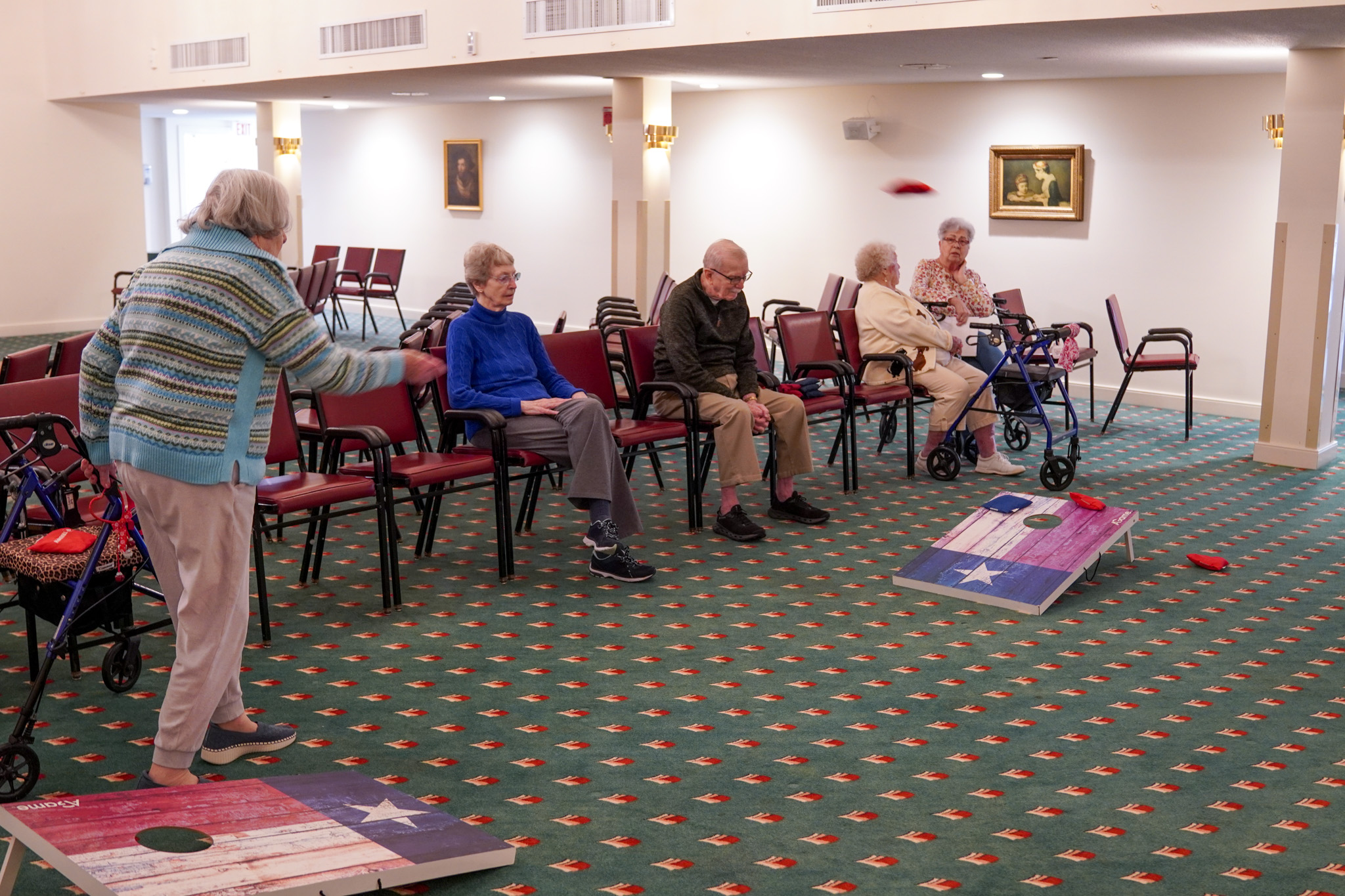 Elderly people, embracing the simplicity that comes with downsizing, enjoy a lively game of indoor bean bag toss in a cozy community room filled with chairs.