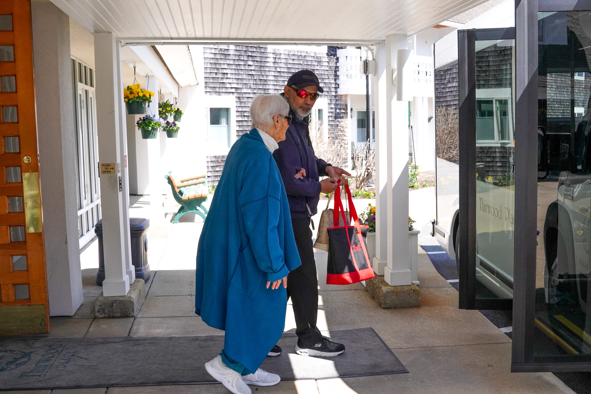 An older woman in a blue coat walks with a man in a cap holding red bags, heading towards a vehicle at an entrance.
