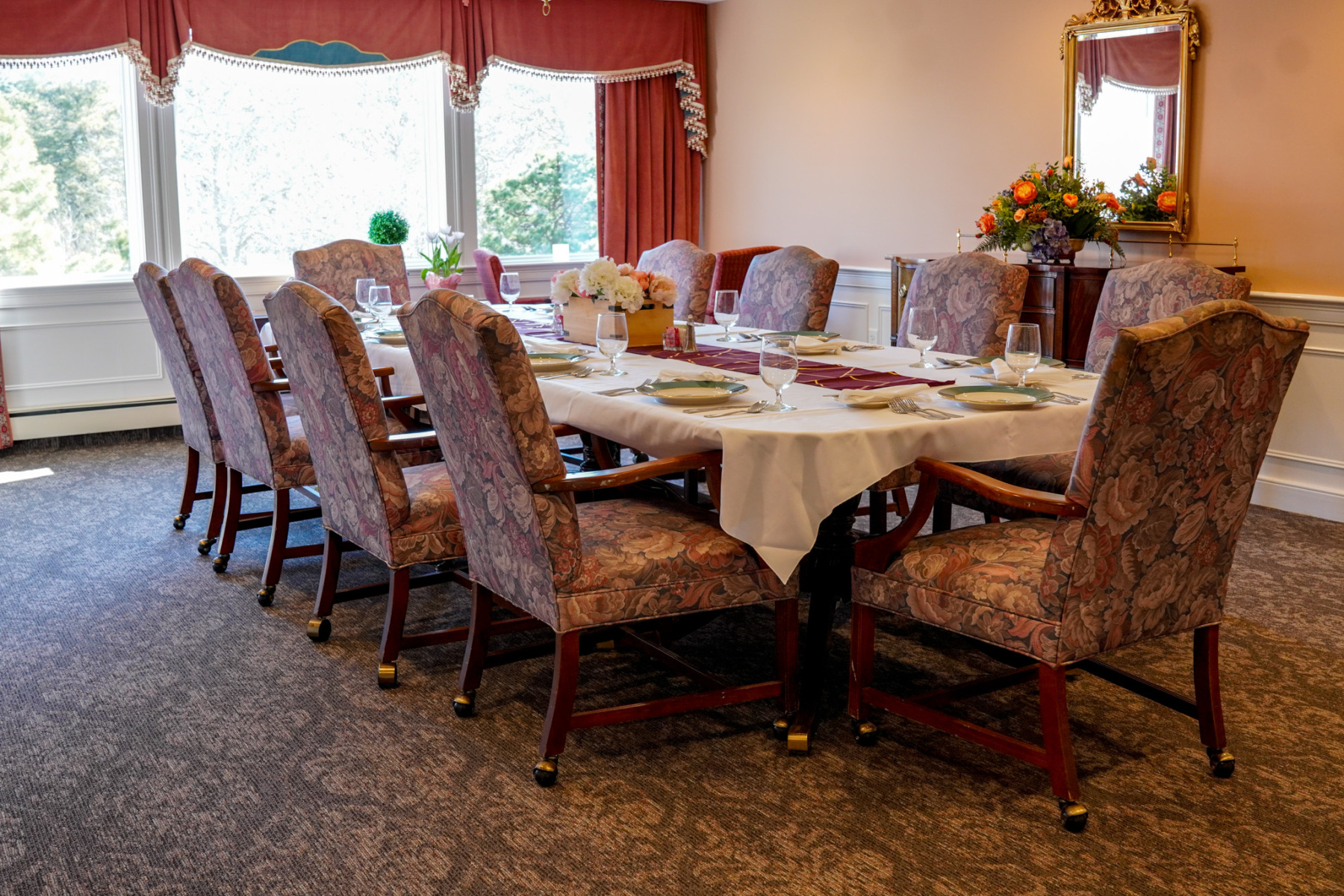 A formal dining table set for eight with floral chairs in an elegant, sunlit room.