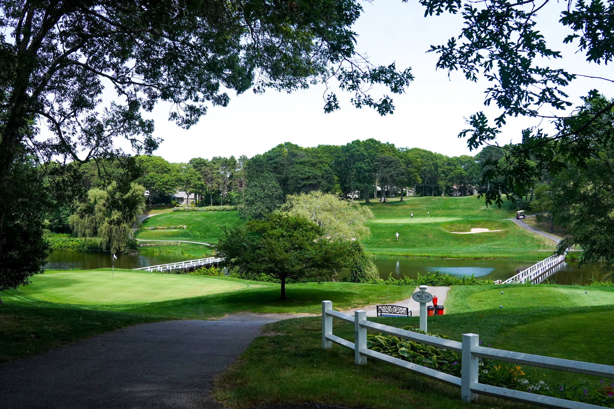 A scenic golf course with green fairways, a pond, white bridges, and trees under a clear sky—perfect for golfers and walkers alike to enjoy the peaceful surroundings.