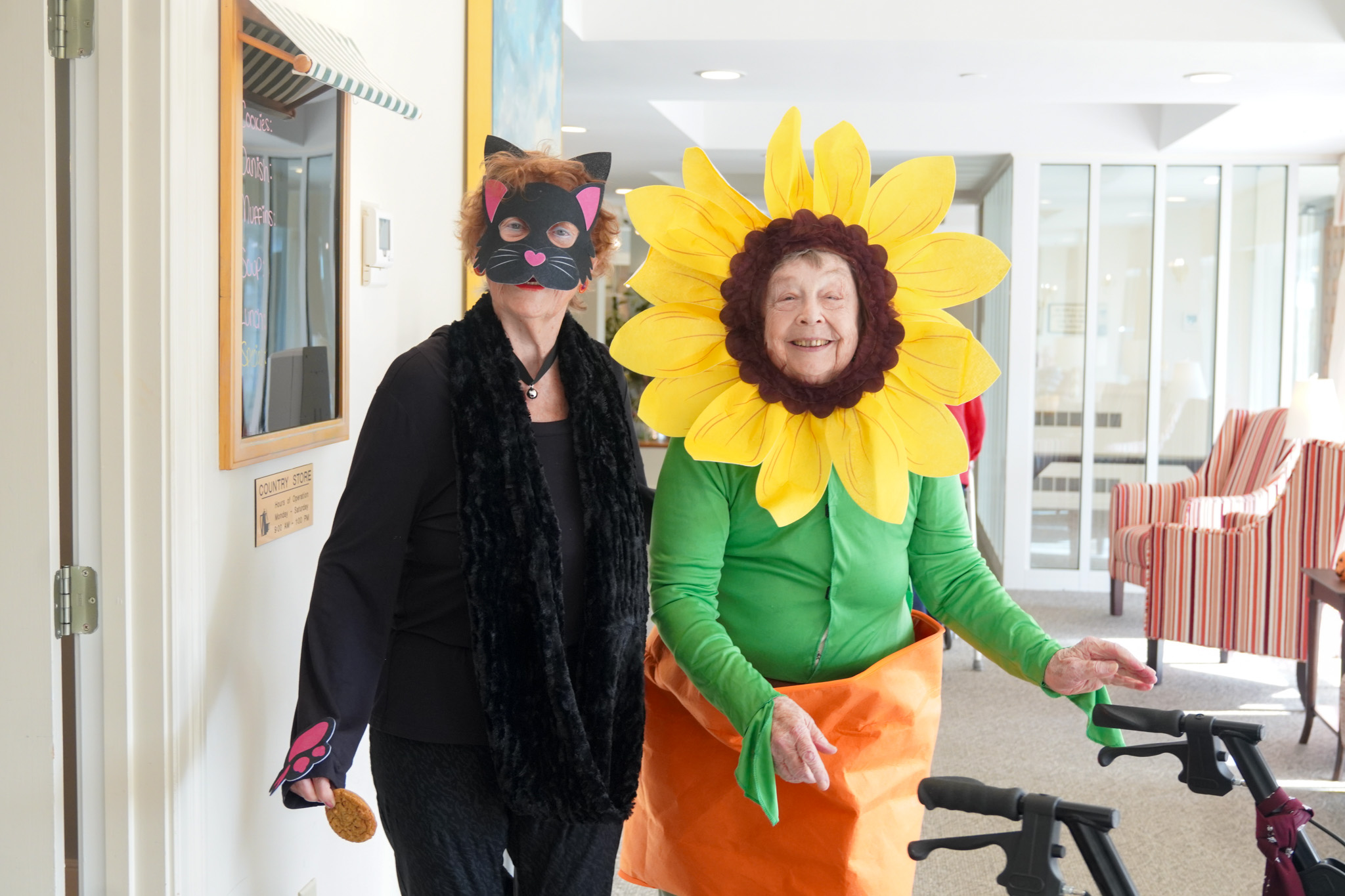 Two elderly women in costumes, one as a black cat and the other as a sunflower, share warm smiles indoors—celebrating friendship and the joy that comes with years of wisdom and scholarship.