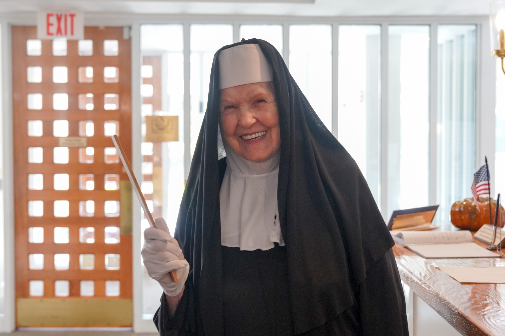 A smiling nun in black and white habit, embodying the spirit of scholarship, holds a ruler while standing in a brightly lit room.