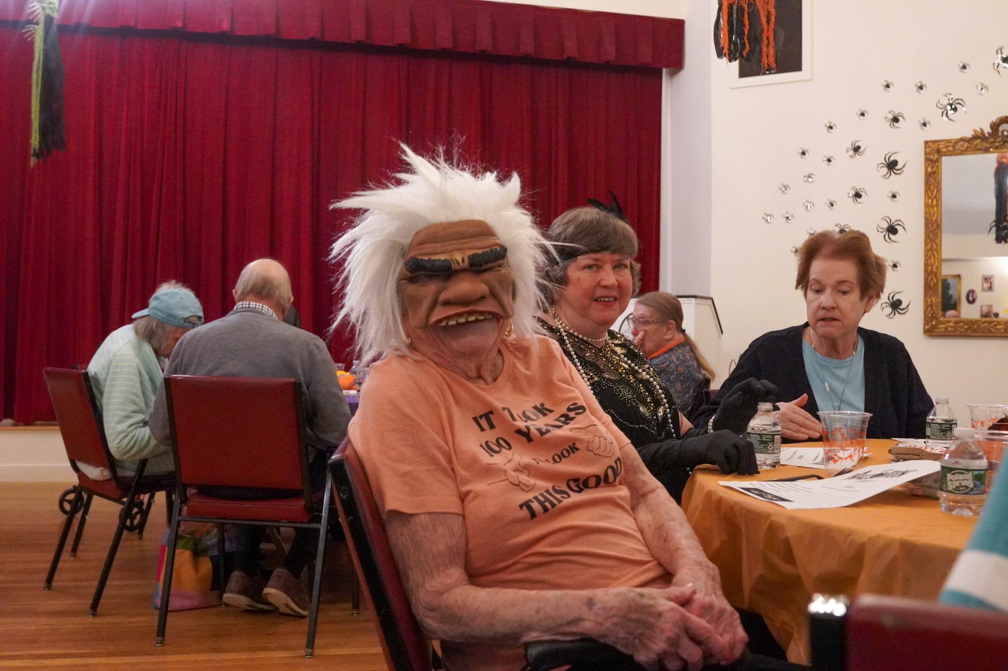 An elderly person in a wild mask sits at a decorated table with others during a festive gathering, sharing stories of scholarship and wisdom.