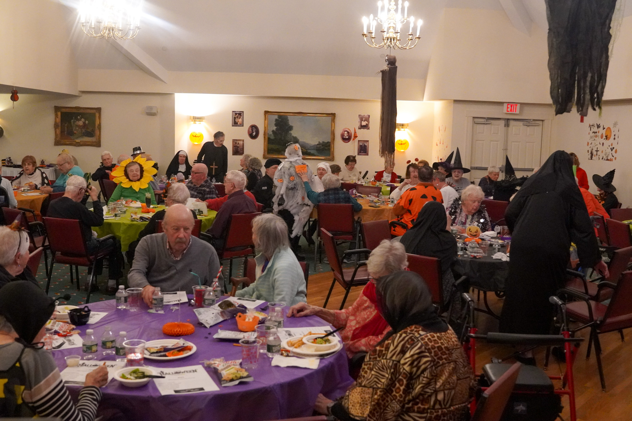 Elderly people in costumes enjoy a Halloween party, eating and socializing in a decorated hall where stories of scholarship and lifelong learning are shared among friends.