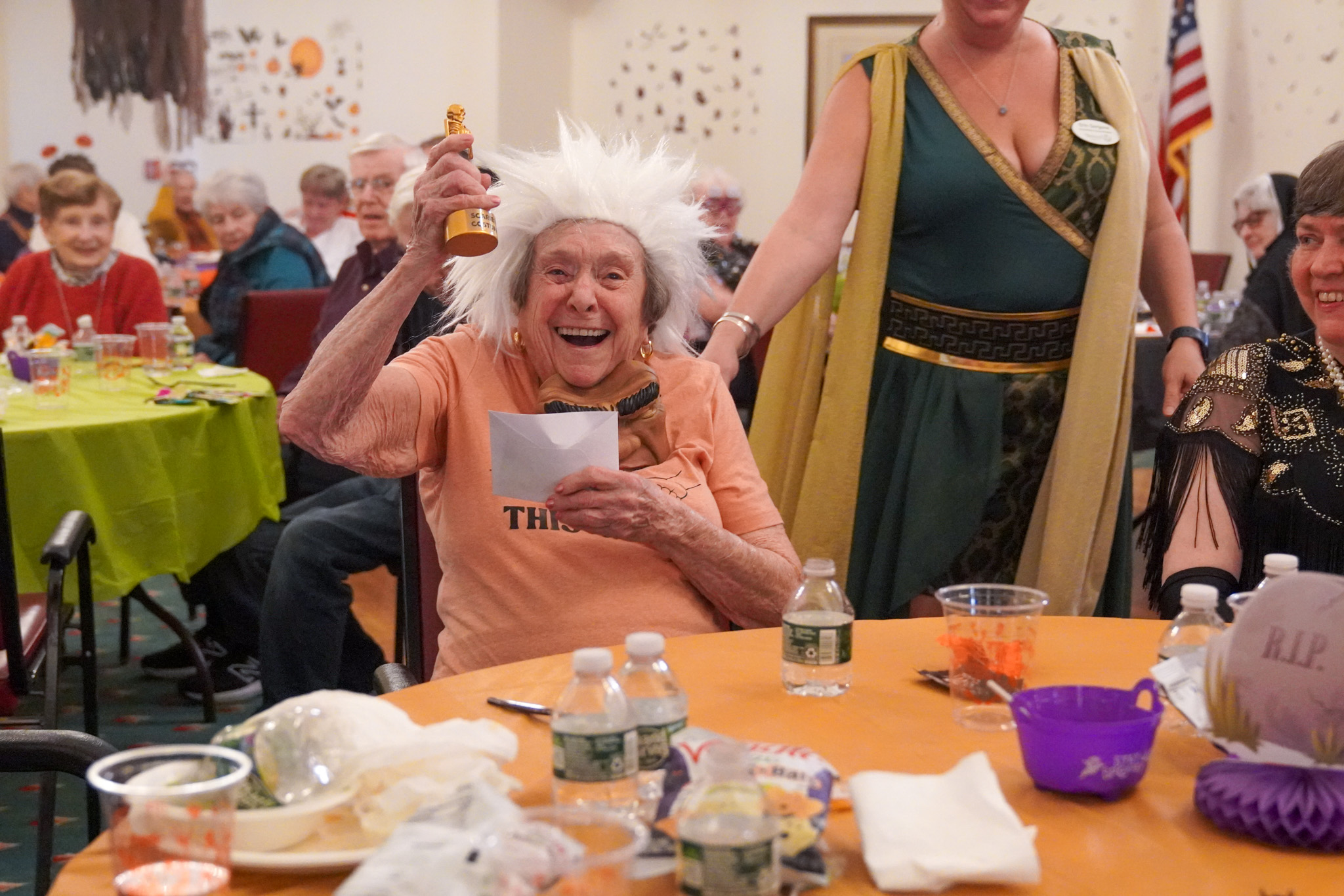Elderly woman in a white wig excitedly holds up a scholarship trophy at a festive party with others in costumes.