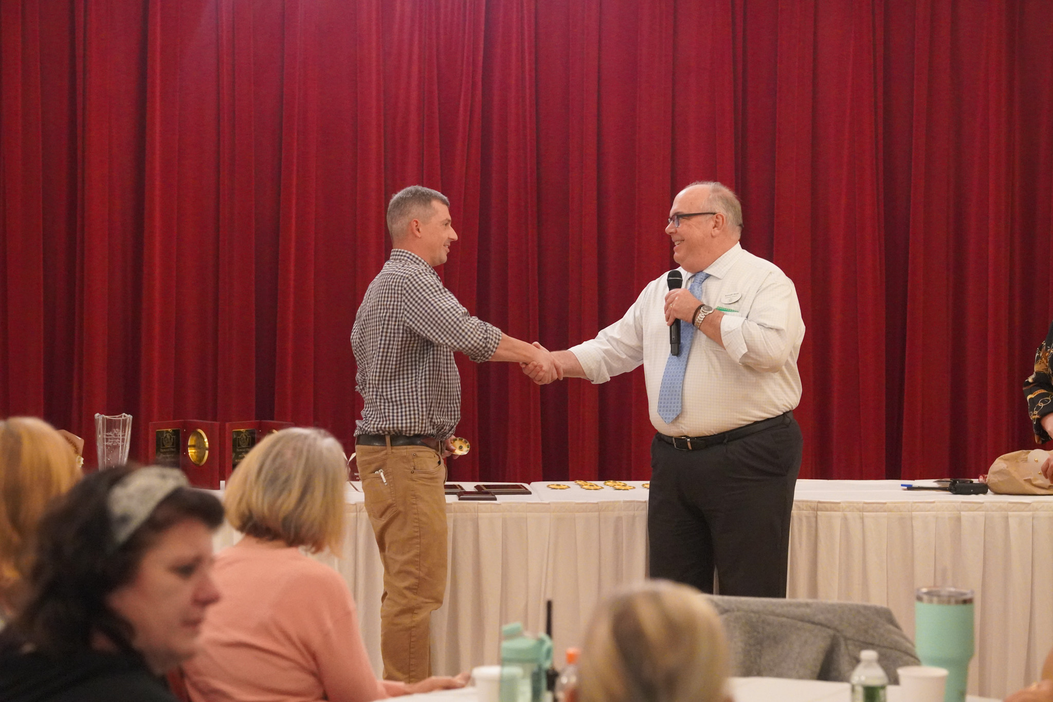 Two men shake hands on stage in front of a red curtain, with people seated and watching.