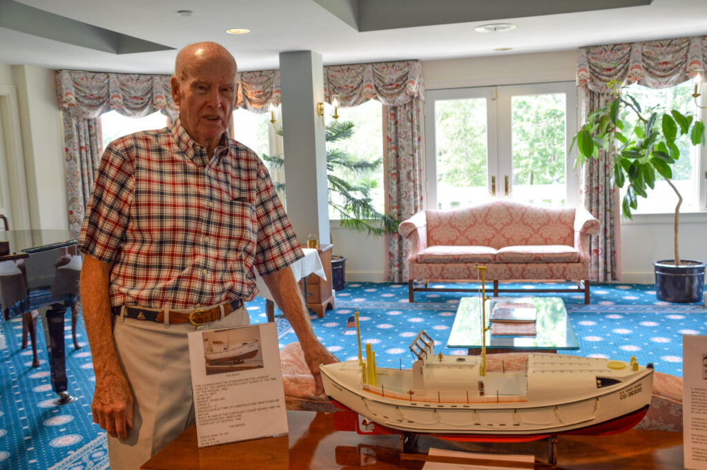 In a bright, elegantly furnished room, an elderly man, embodying the spirit of senior living, stands proudly next to a detailed model boat resting on the table.