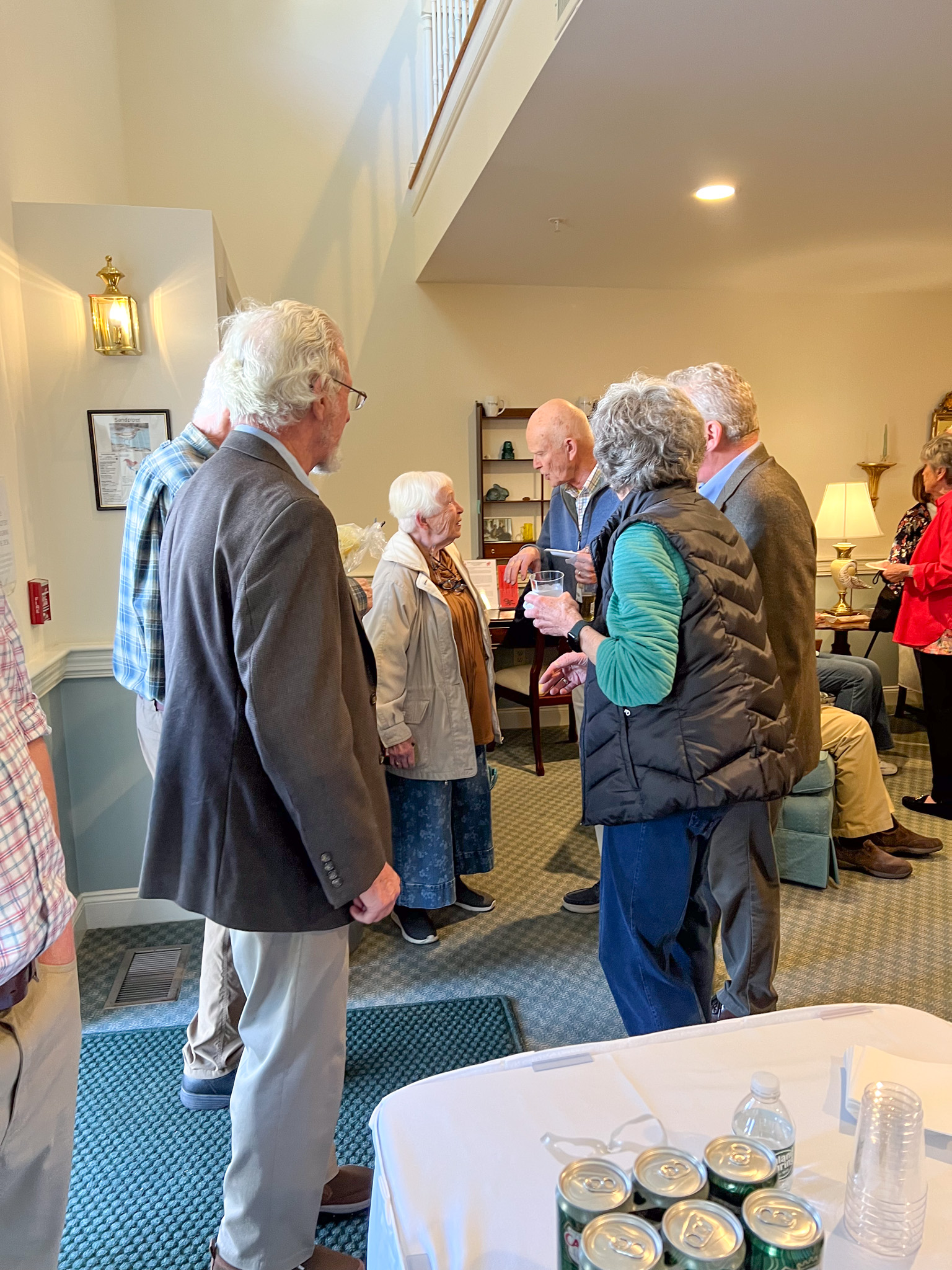 A group of older adults talking and socializing indoors at a casual gathering, sharing stories about family, travel, and scholarship experiences.