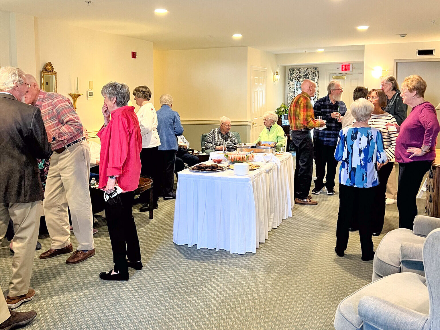 A group of older adults, some sharing stories about scholarship opportunities, socializing and serving food at a gathering in a bright, carpeted room.