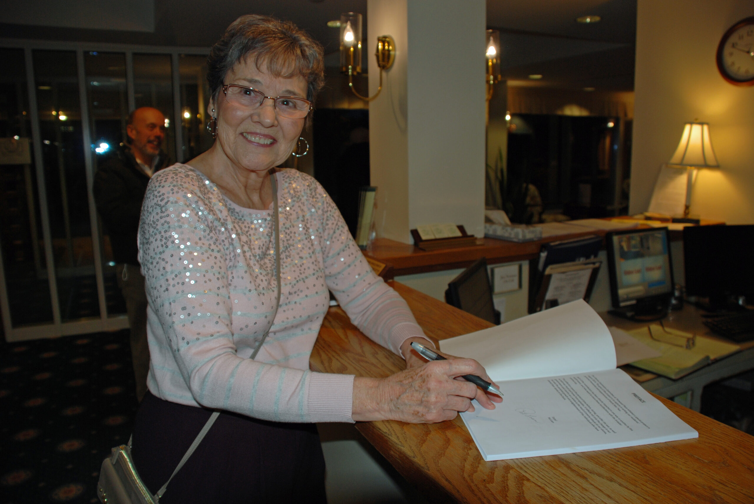 An older woman, beaming with joy, signs a document at a wooden counter indoors, each stroke of the pen telling countless stories of her journey.