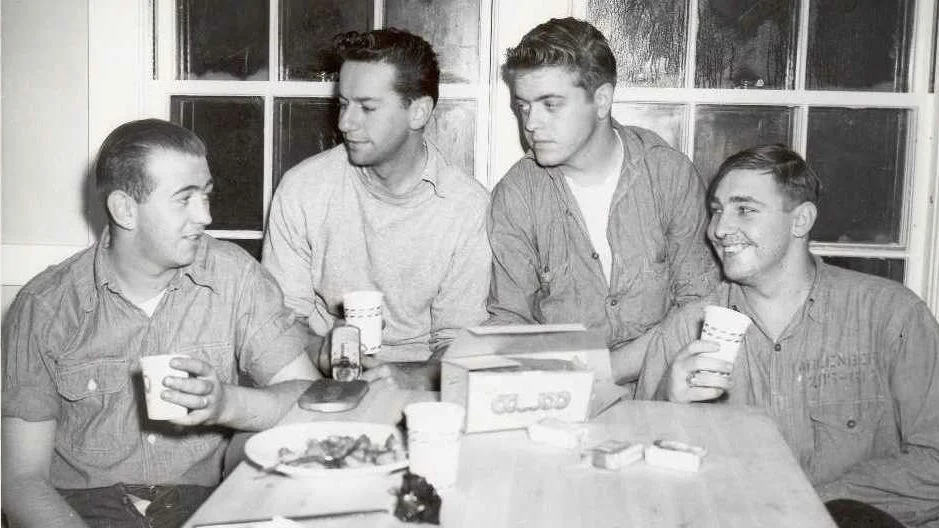 Four men sitting at a table with drinks and snacks, smiling and talking as casually as sailors swapping stories aboard ships.