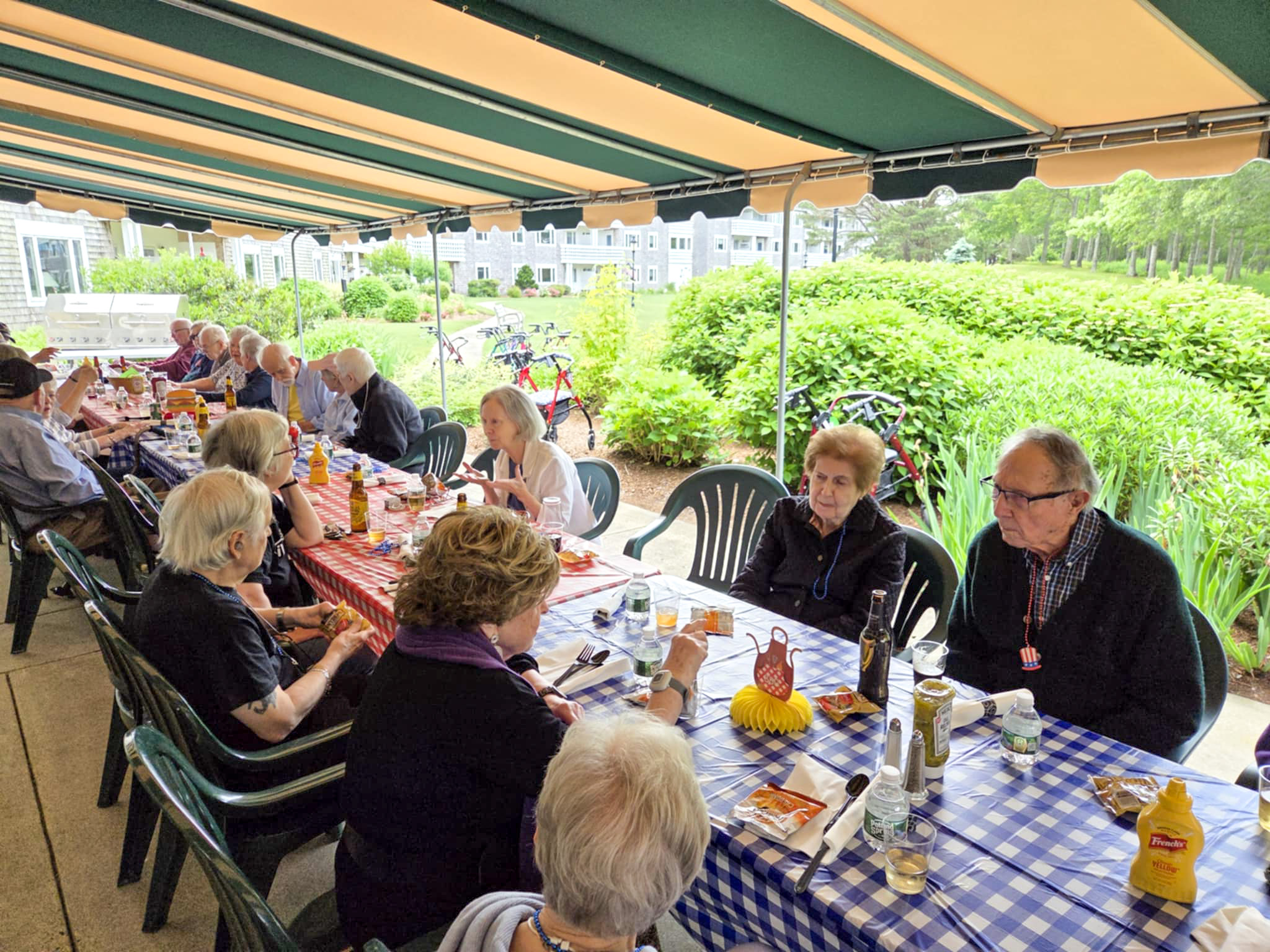 A group of seniors sit at long picnic tables under an awning, enjoying a summer meal together outdoors.