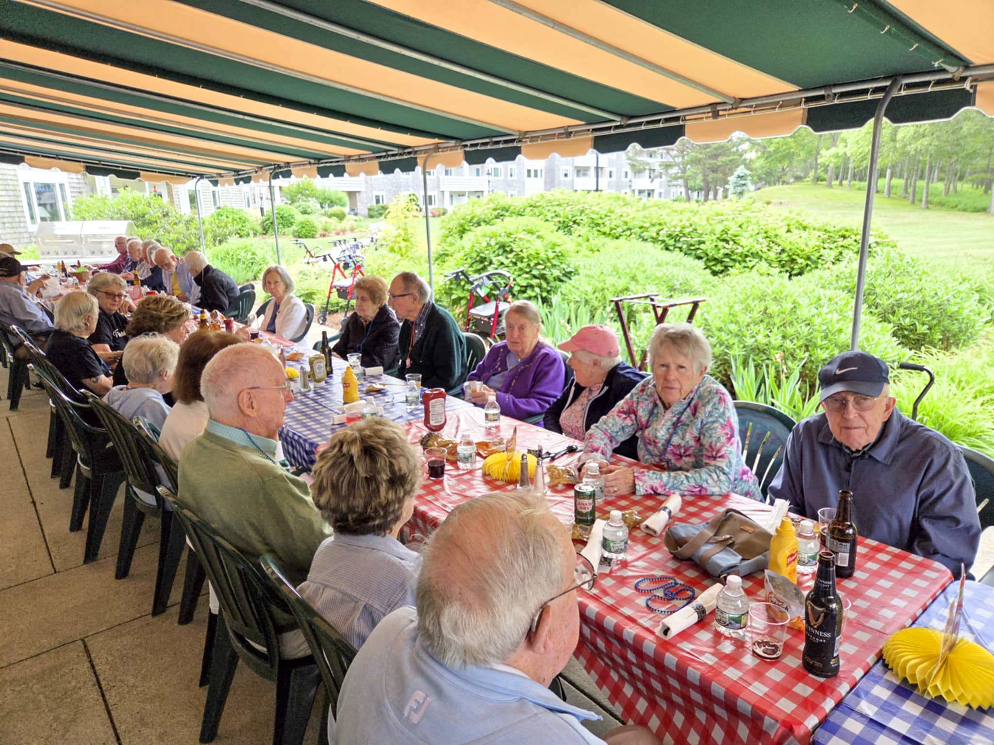 A group of seniors sit at long picnic tables outdoors, enjoying a summer meal under an awning.