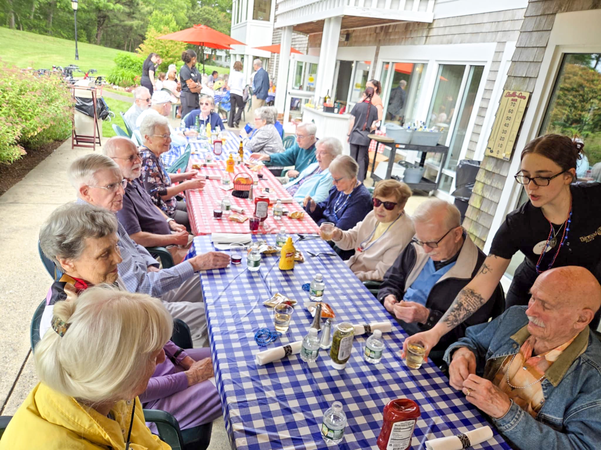 A group of seniors enjoys a summer gathering, sitting at a long outdoor table with checkered tablecloths, being served food and drinks.