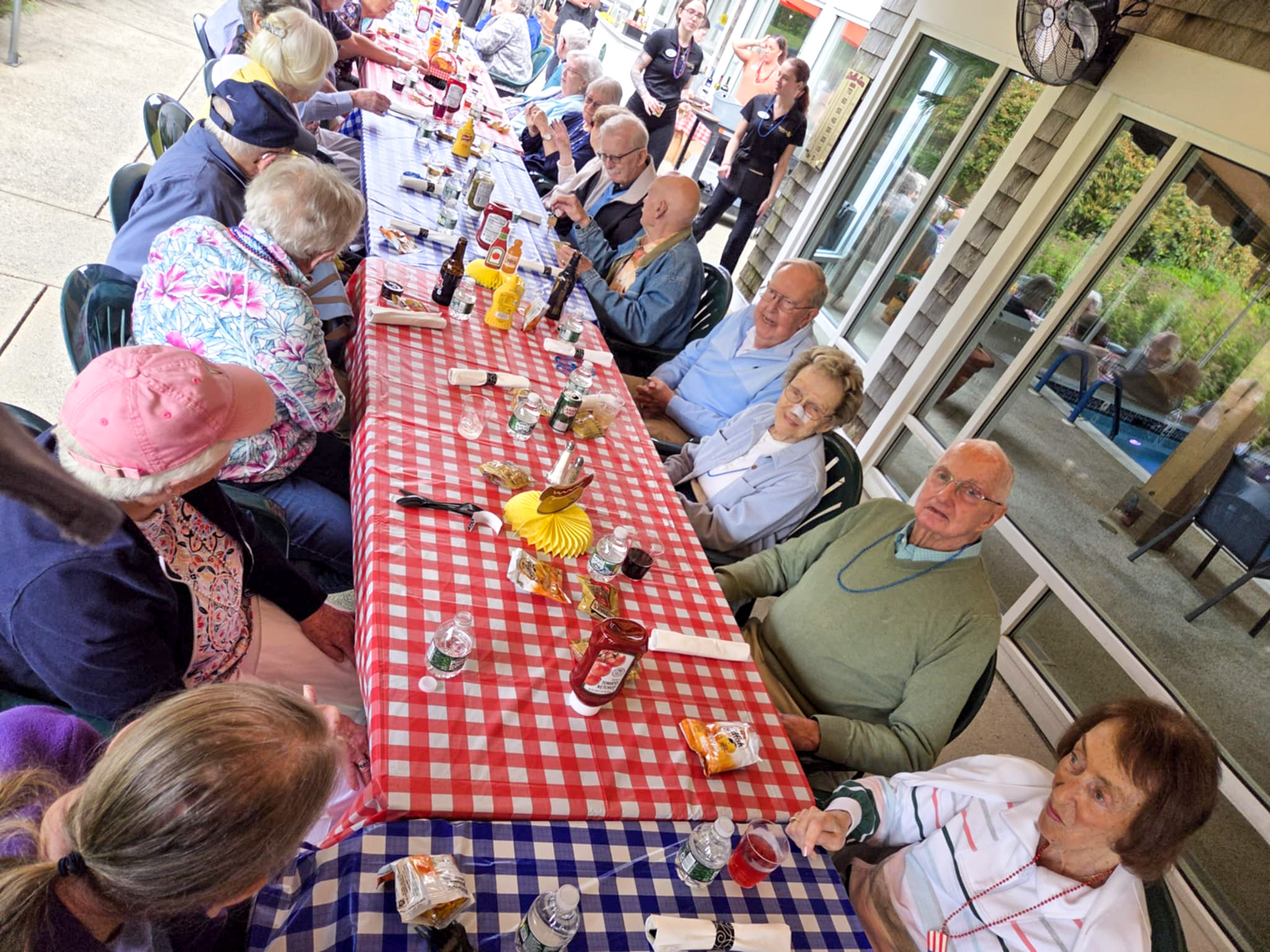 A group of seniors sit around tables with red and blue checkered tablecloths at a cheerful summer outdoor gathering.