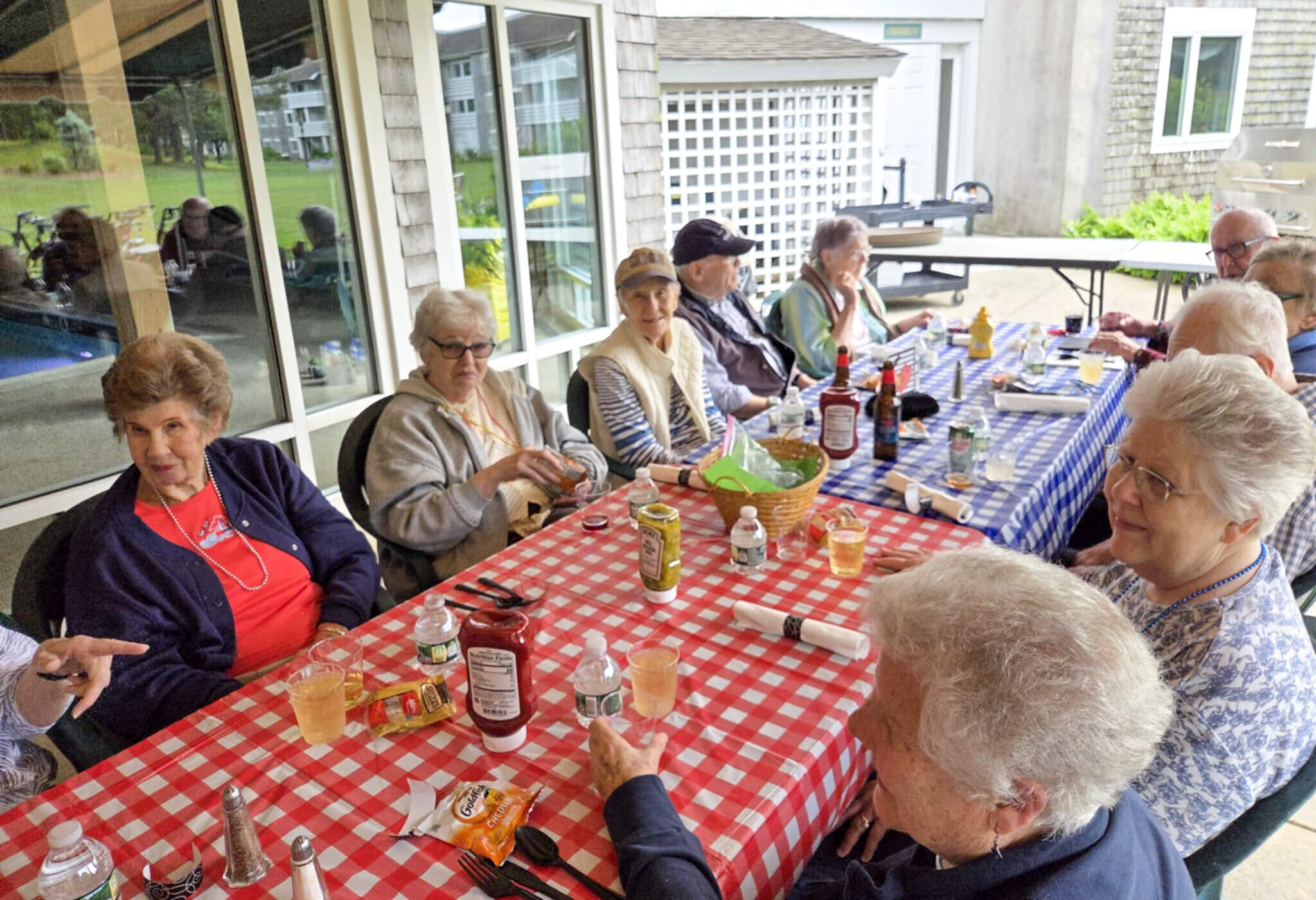 A group of seniors sit around picnic tables with drinks and food, enjoying a relaxing summer outdoor gathering.