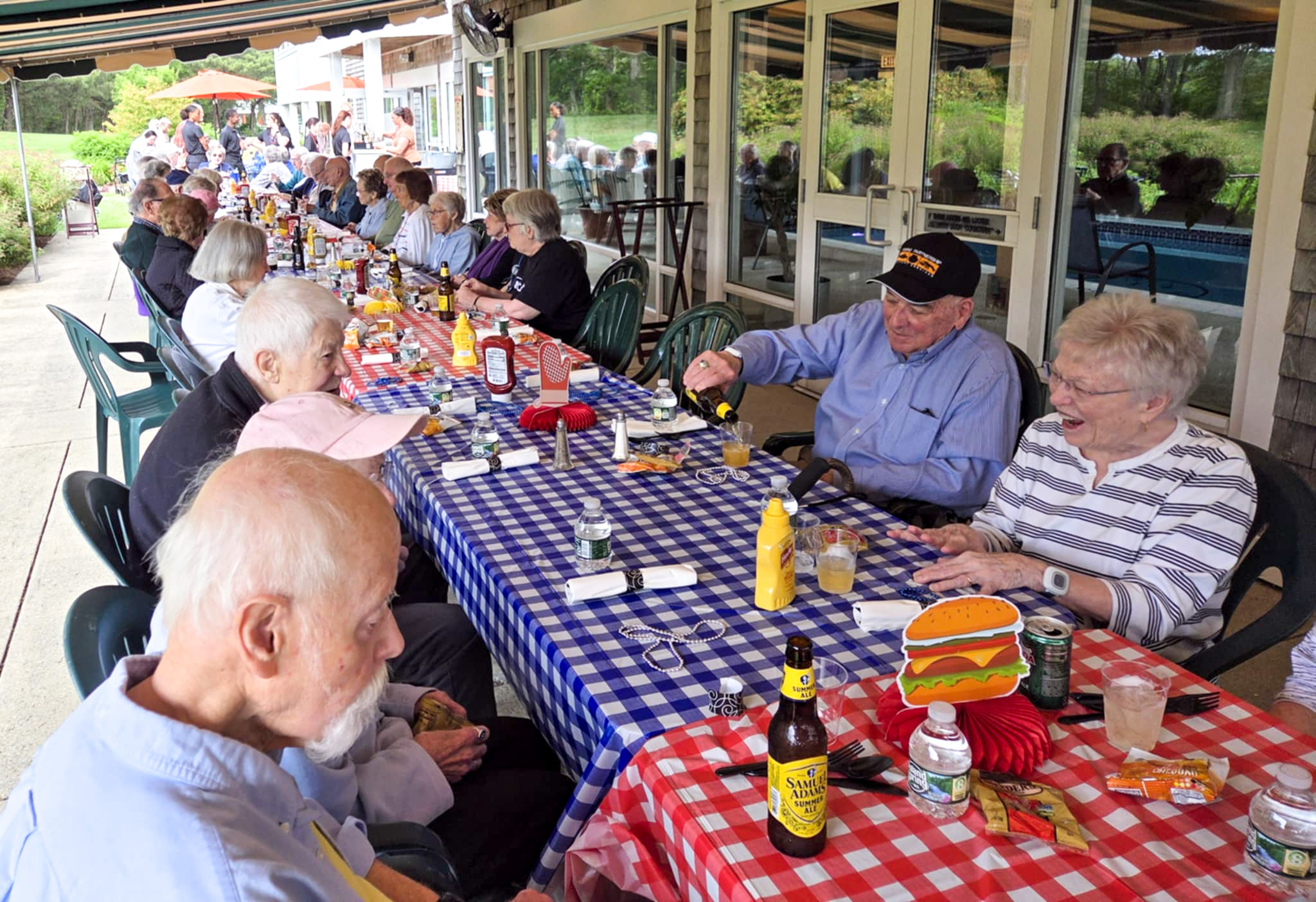A group of seniors enjoy a summer picnic meal together at long tables covered with red and blue checkered cloths.
