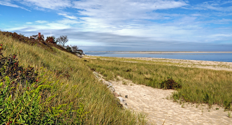 Grassy dunes lead to a serene sandy beach and calm blue sea under a partly cloudy sky, offering an ideal setting for senior living.