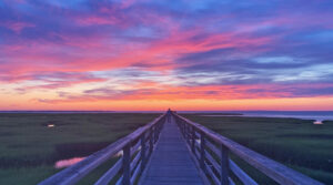 A wooden boardwalk leads to the horizon at sunset, where vibrant purple, pink, and orange clouds glow overhead like a skyborne lightship.