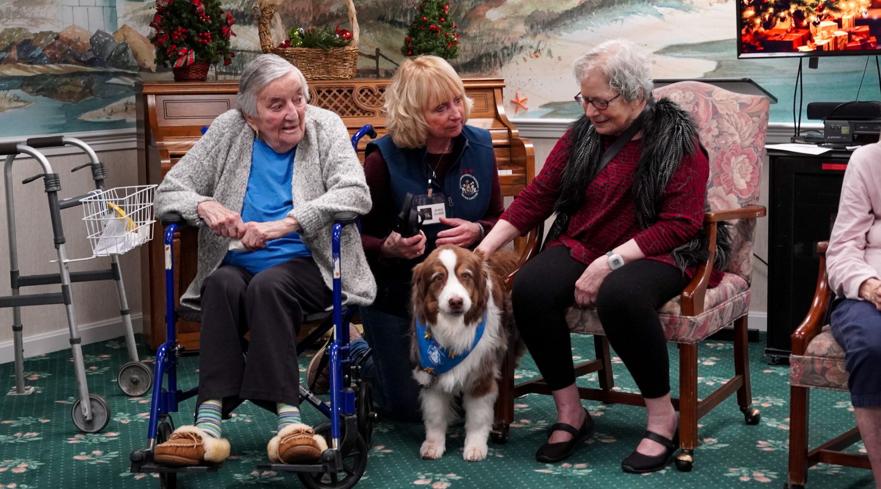 Two elderly women and a caregiver sit indoors with an animal therapy dog, smiling and petting the dog.