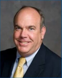 Smiling middle-aged man in a suit and yellow tie, exuding confidence against a gray background.