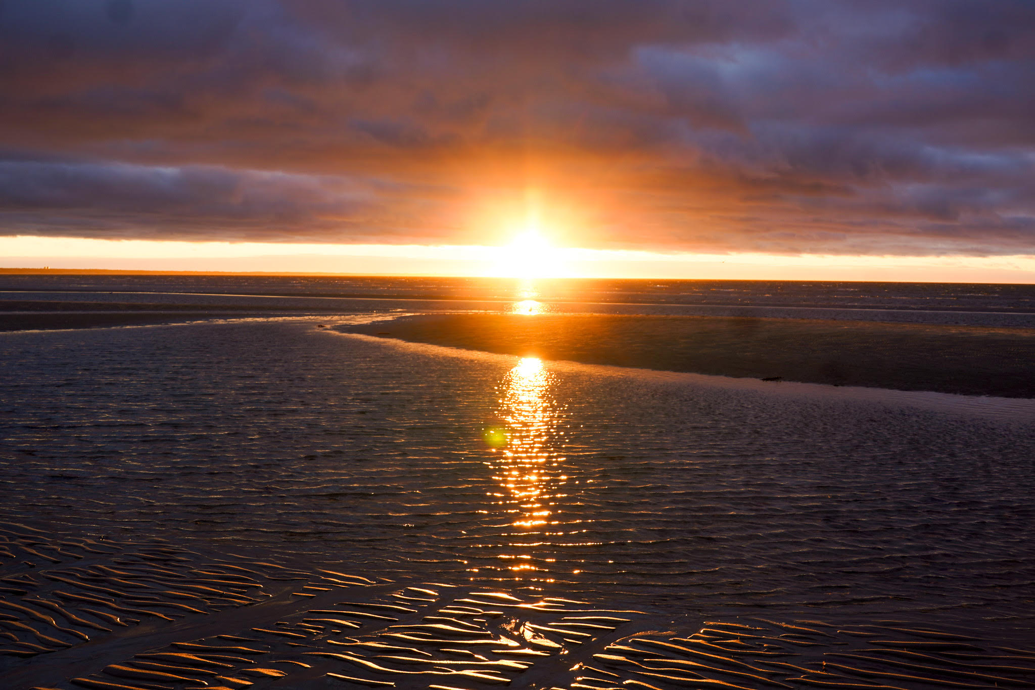 Sunset over a beach with rippled sand and water reflecting the golden light, creating a serene backdrop reminiscent of tranquil senior living. Dark clouds partially obscure the horizon, adding an air of mystery.