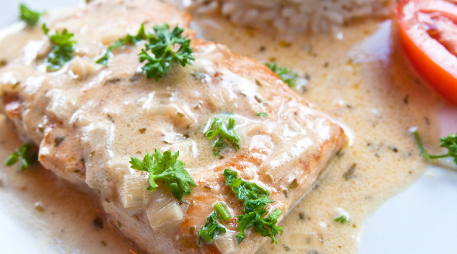 Close-up of a salmon fillet with creamy sauce, garnished with parsley. Served with rice and a slice of tomato on a white plate.