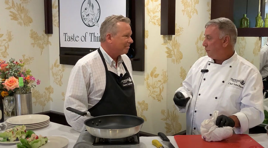 Two chefs talk in a kitchen studio, one in a white coat, the other in a black apron, with food and flowers nearby.