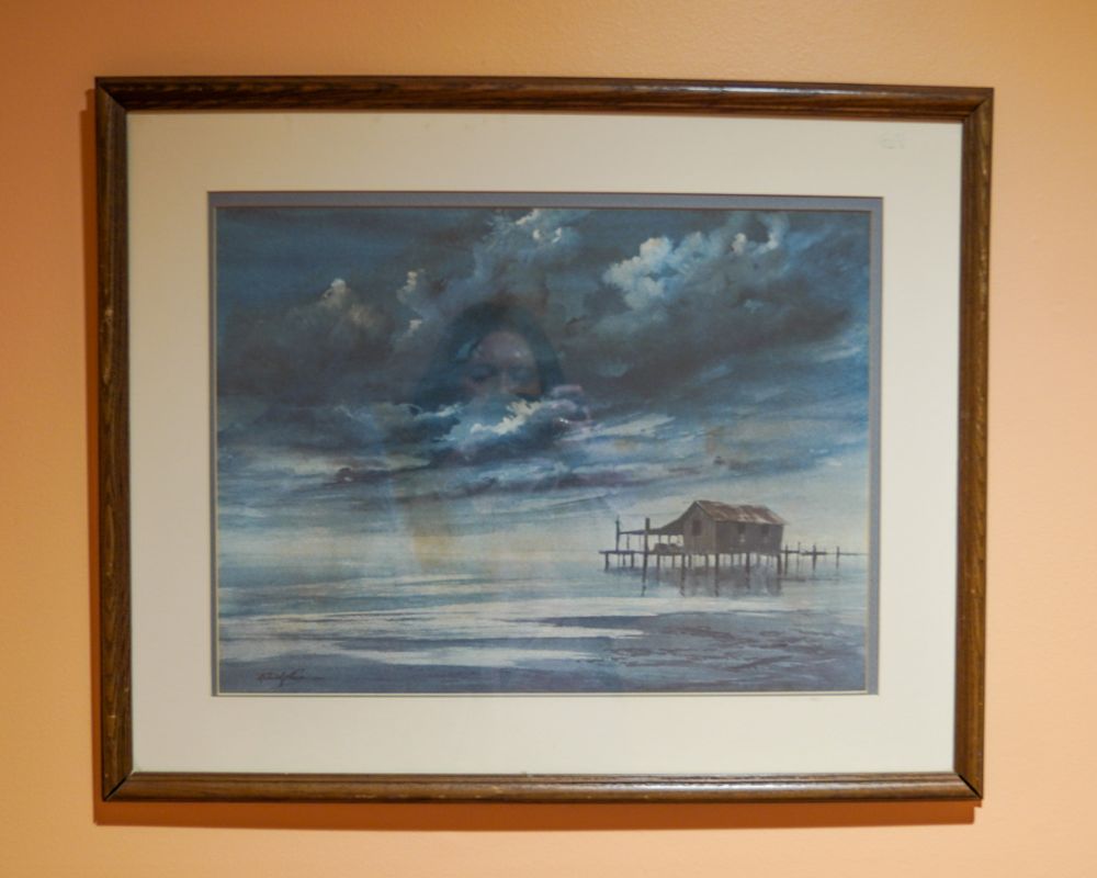 Framed painting of a pier and house over water, under a cloudy sky, evokes tranquility, much like the peacefulness found in senior living. A reflected image of a person is visible on the glass, adding depth to the scene.